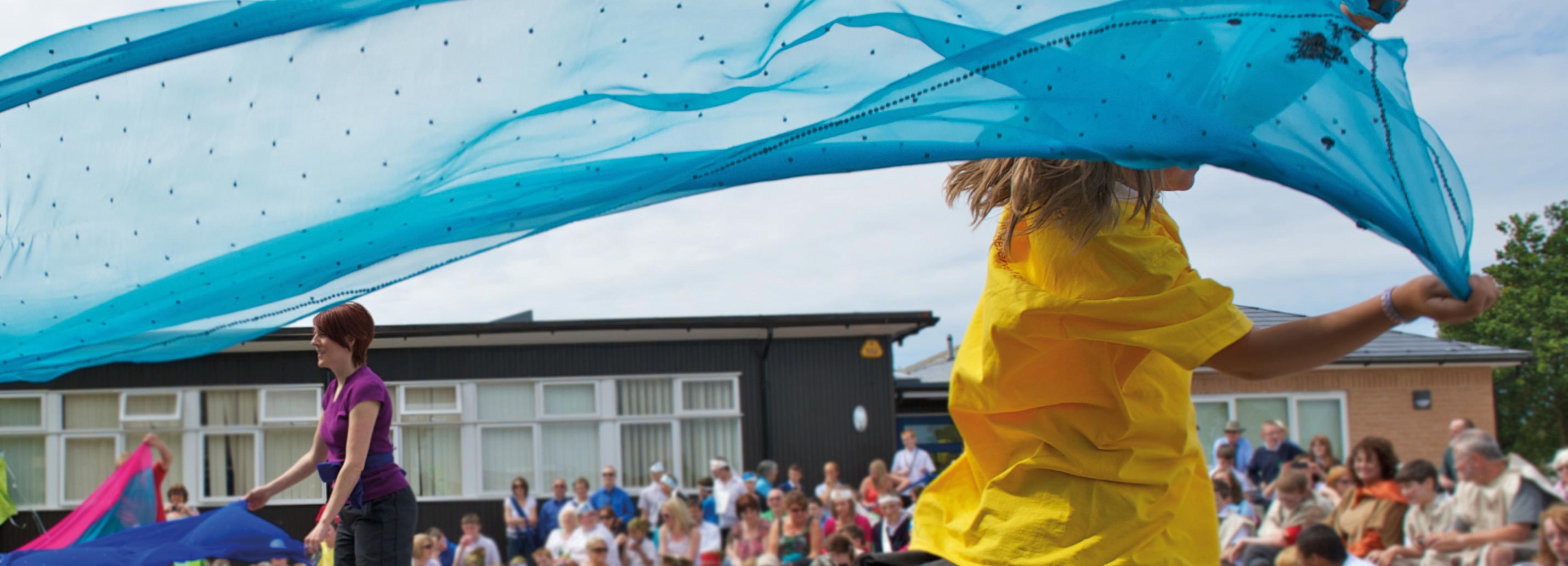 A young girl running with a ribbon on a primary school playground with teachers, parents and pupils eagerly sat watching.