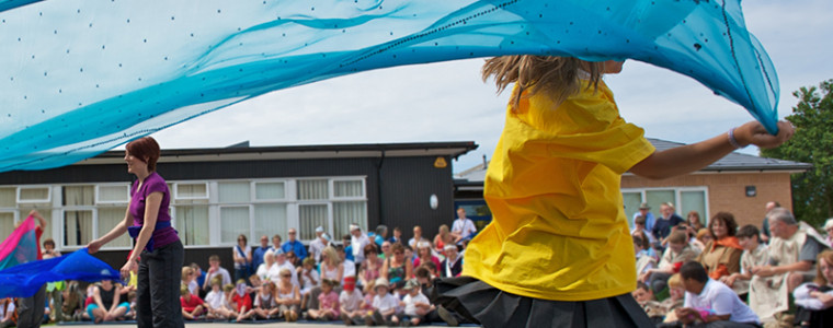 A young girl running with a ribbon on a primary school playground with teachers, parents and pupils eagerly sat watching.