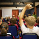 School boy in the foreground with his arm raised in a classroom full of children all sat facing an electronic white board.