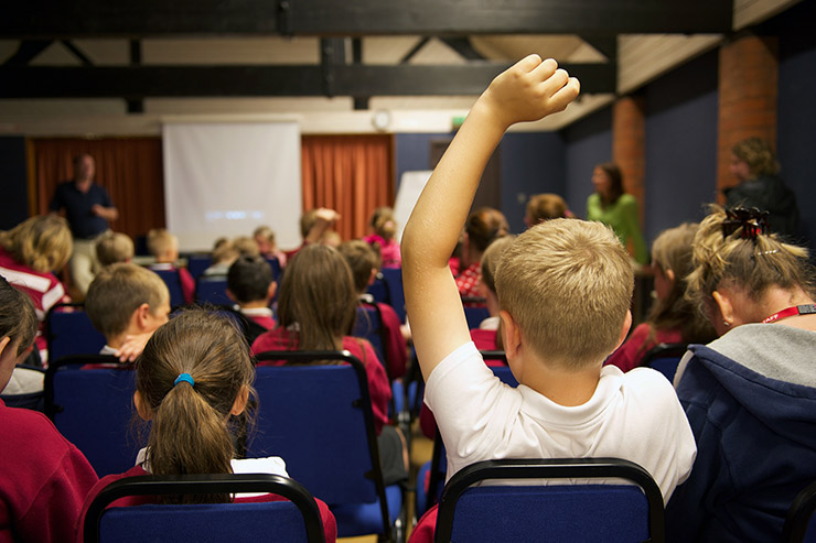 School boy in the foreground with his arm raised in a classroom full of children all sat facing an electronic white board.