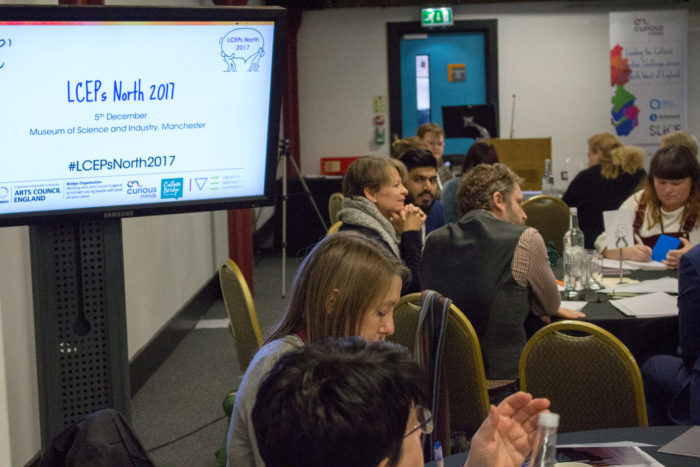 A diverse room of people sat at tables discussing creative ideas. There is a monitor with the writing: "LCEPs North 2017" with the logos of several arts organisations such as Curious Minds and the Arts Council England.