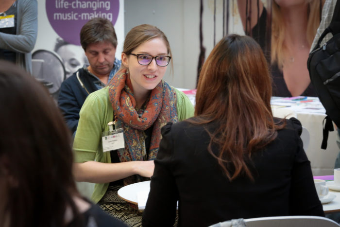 Two women sat down at a table eagerly discussing something of interest in a busy room of people and leaflets.