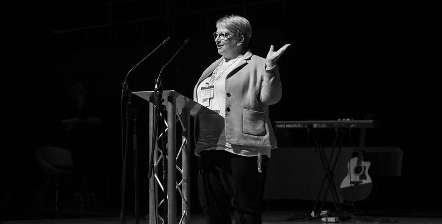 Black and white photo of a smartly dressed woman smiling and presenting on stage.