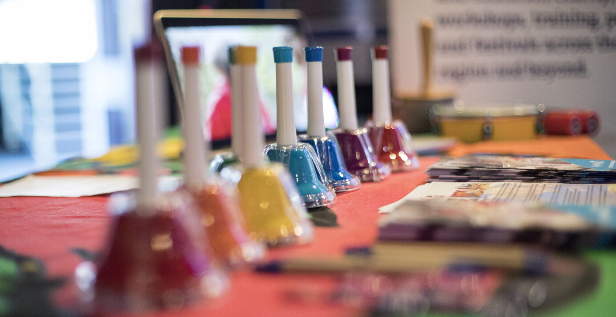 A row of colourful bells laid across a red table with pieces of paper out of focus on the end fo the table.