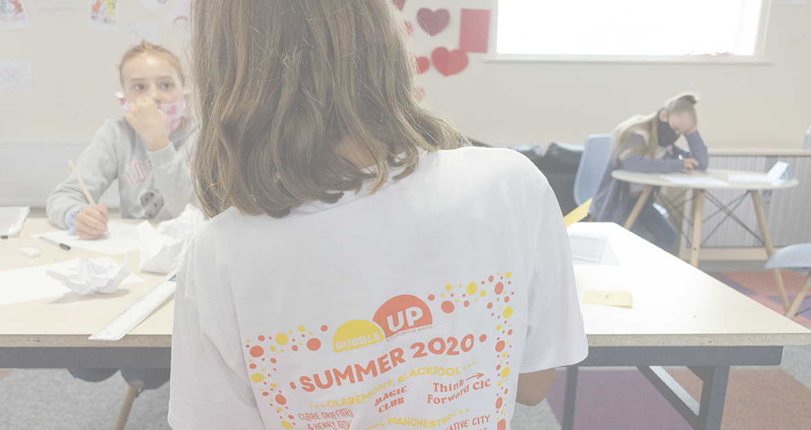 A young Bubble Up participant works at a table with a friend. Both are wearing face coverings. One is wearing a Bubble Up t-shirt.