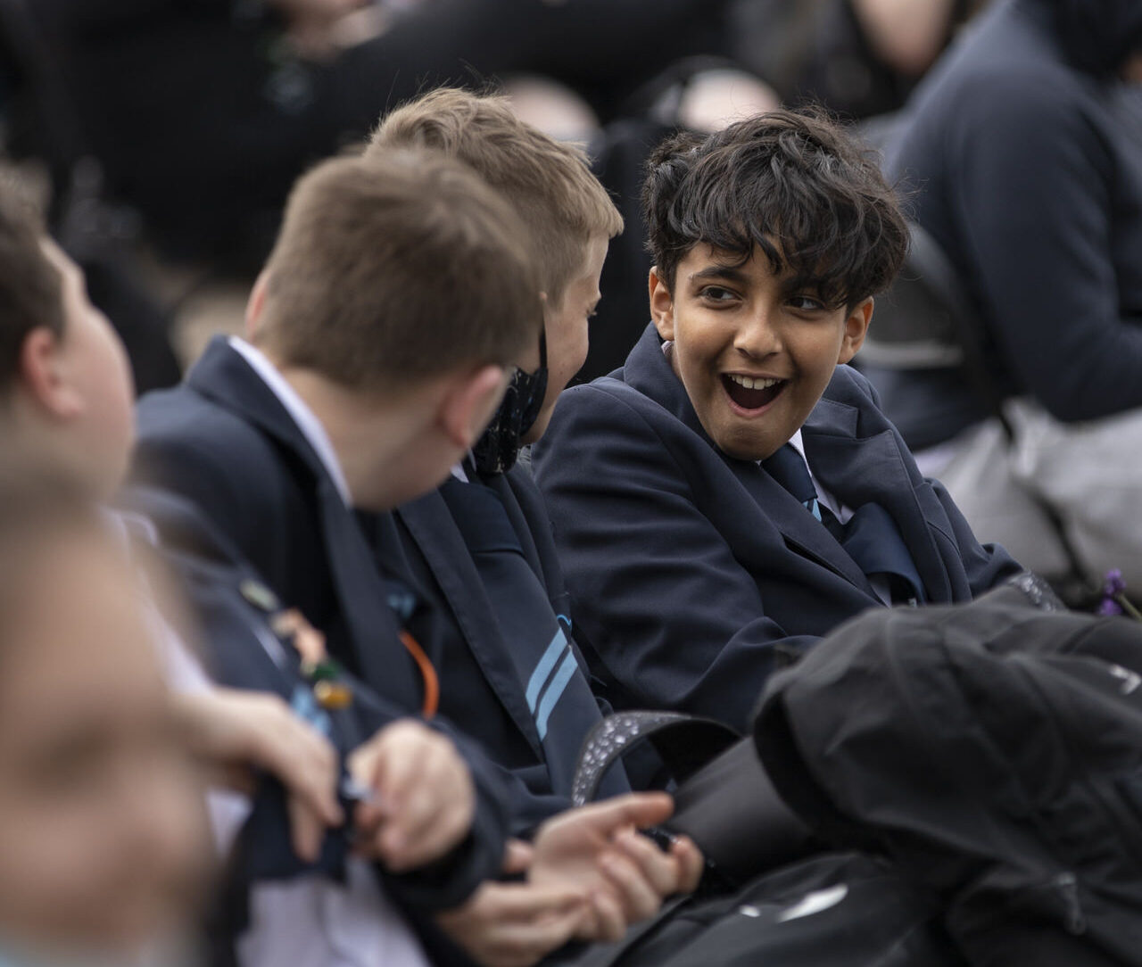 A secondary school pupil in uniform sits with other students as part of the audience at an outdoor show. Their expression is one of excitement, wonder and delight.