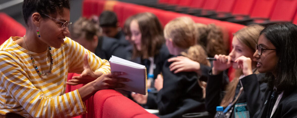 A teacher is with pupils in theatre seating. She is showing them a book and explaining something.