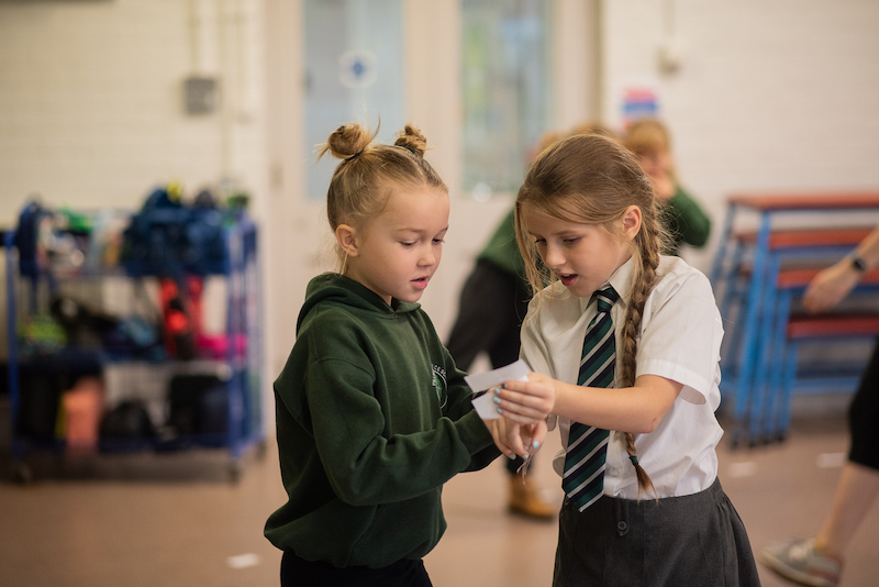 Two primary age children in school uniform interact with each other during an active class. They are concentrating on a piece of paper in one child's hand.
