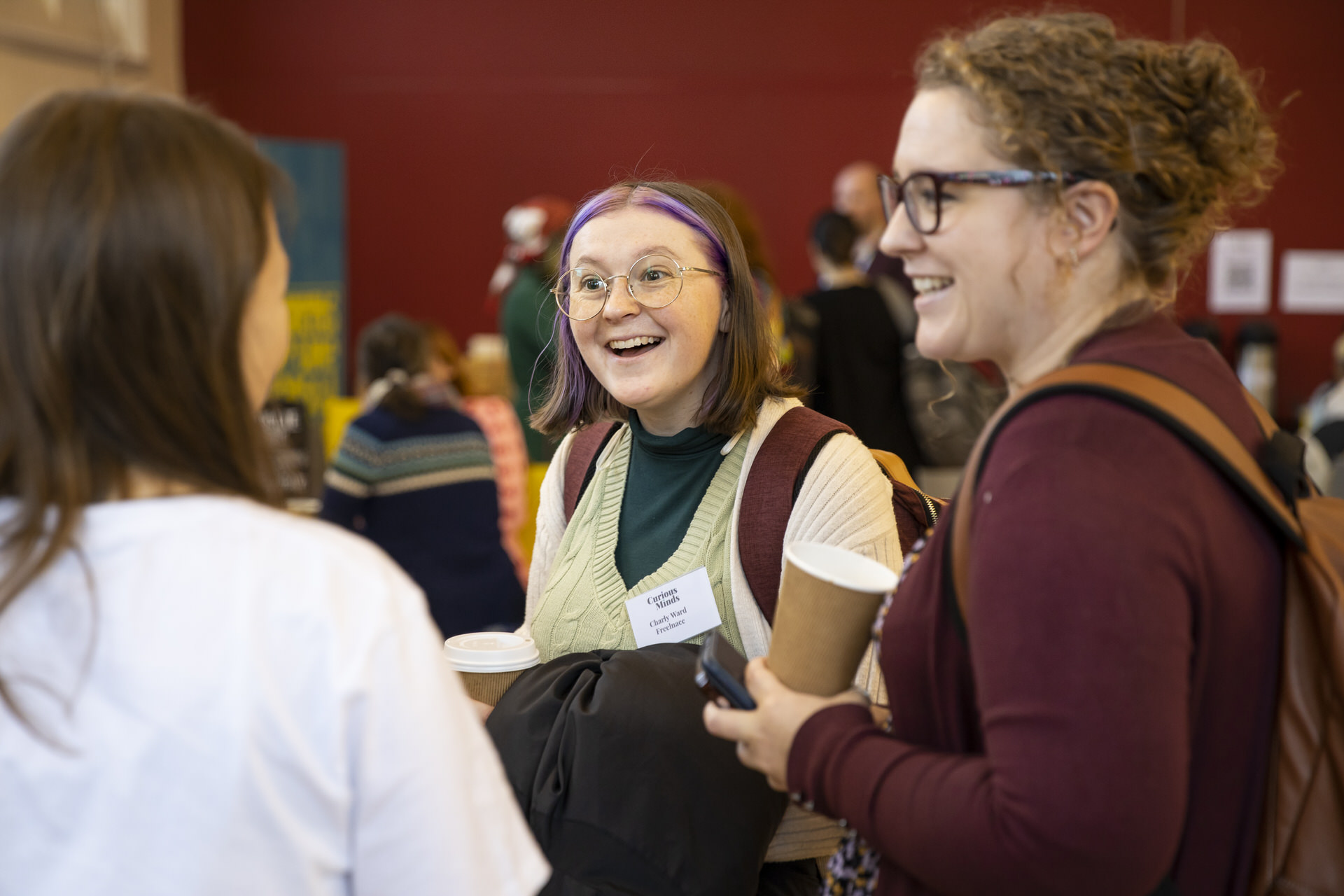 Holly Ball is chatting animatedly with two people at an event. She is wearing a burgundy top and carrying a coffee cup.