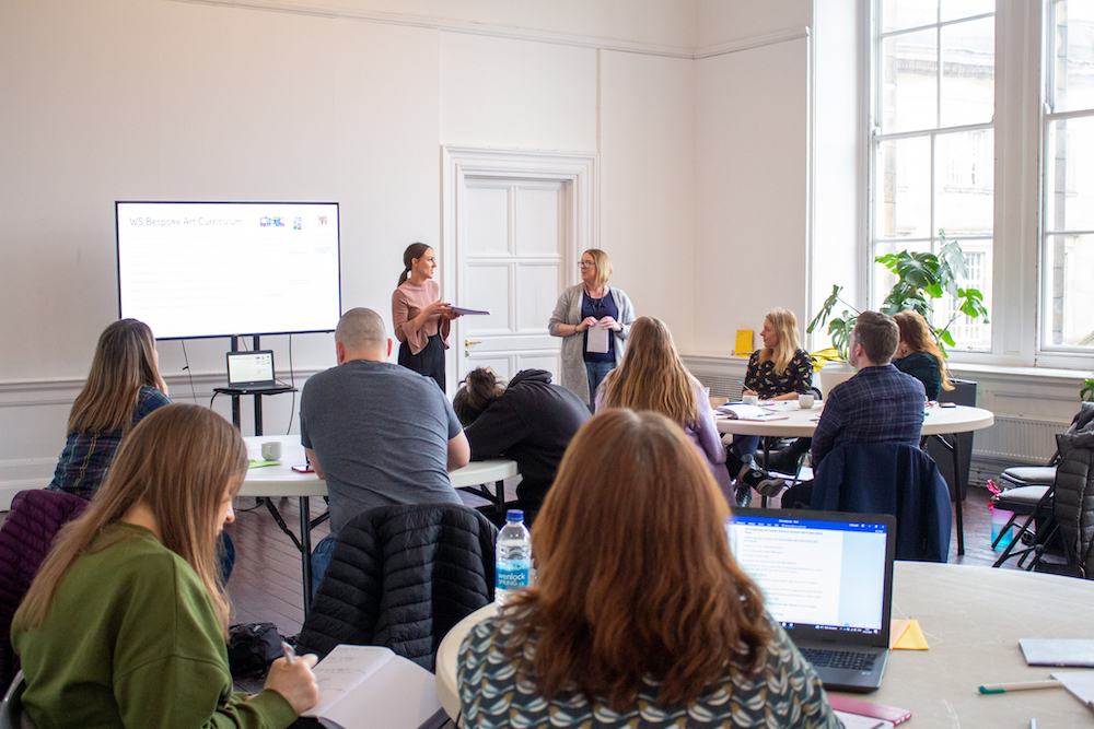 Leading the Arts in your School delegates at a training day. Participants are sat at tables in a light, airy room. They are listening to a trainer facilitator who is speaking.