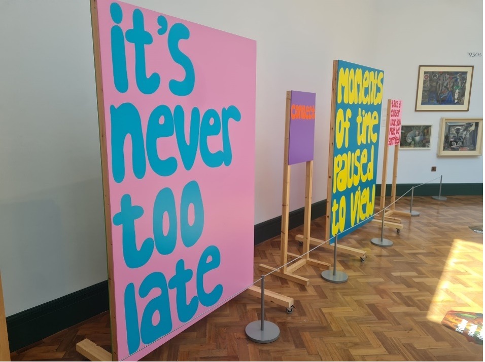 An exhibition with a large artwork that reads "Its never too late" in light blue writing on a pale pink background. Other artworks can be seen in the background.