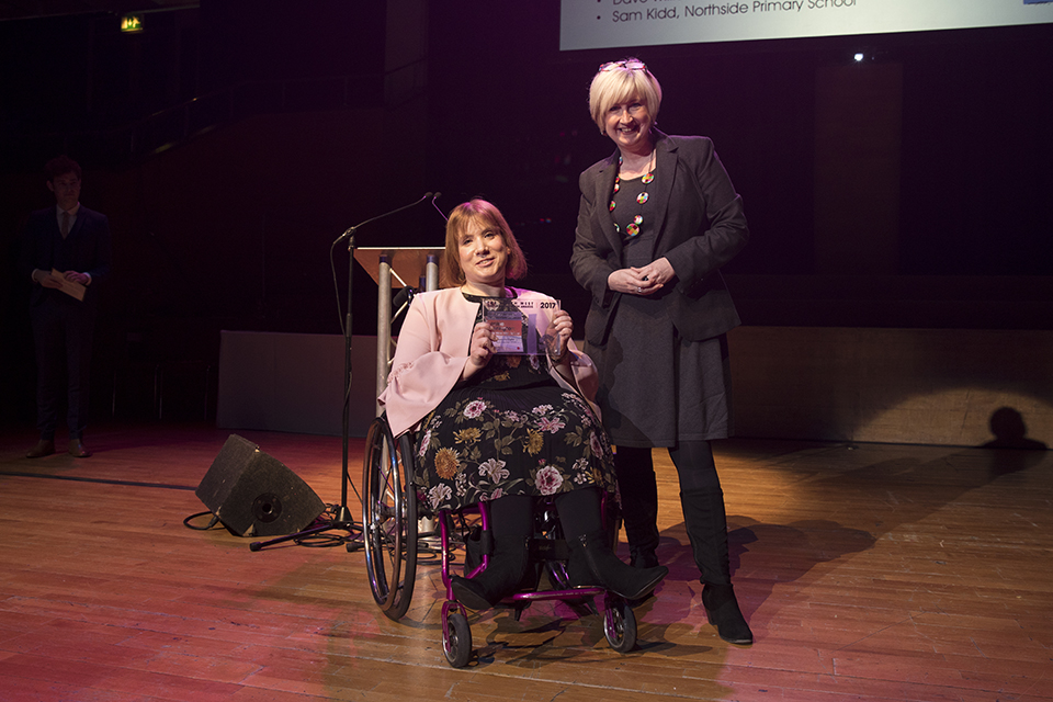 2 women on stage. One of them is in a wheelchair and holding up an award they won.