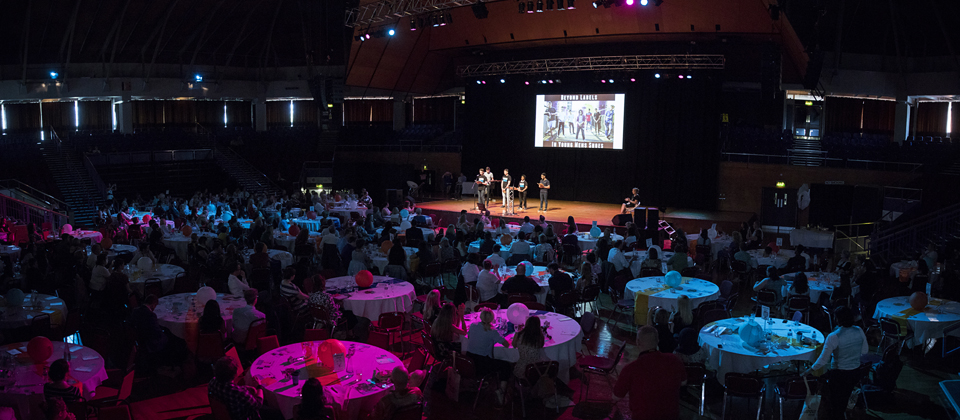 Awards ceremony with stage and a room full of attendees sat round circular tables