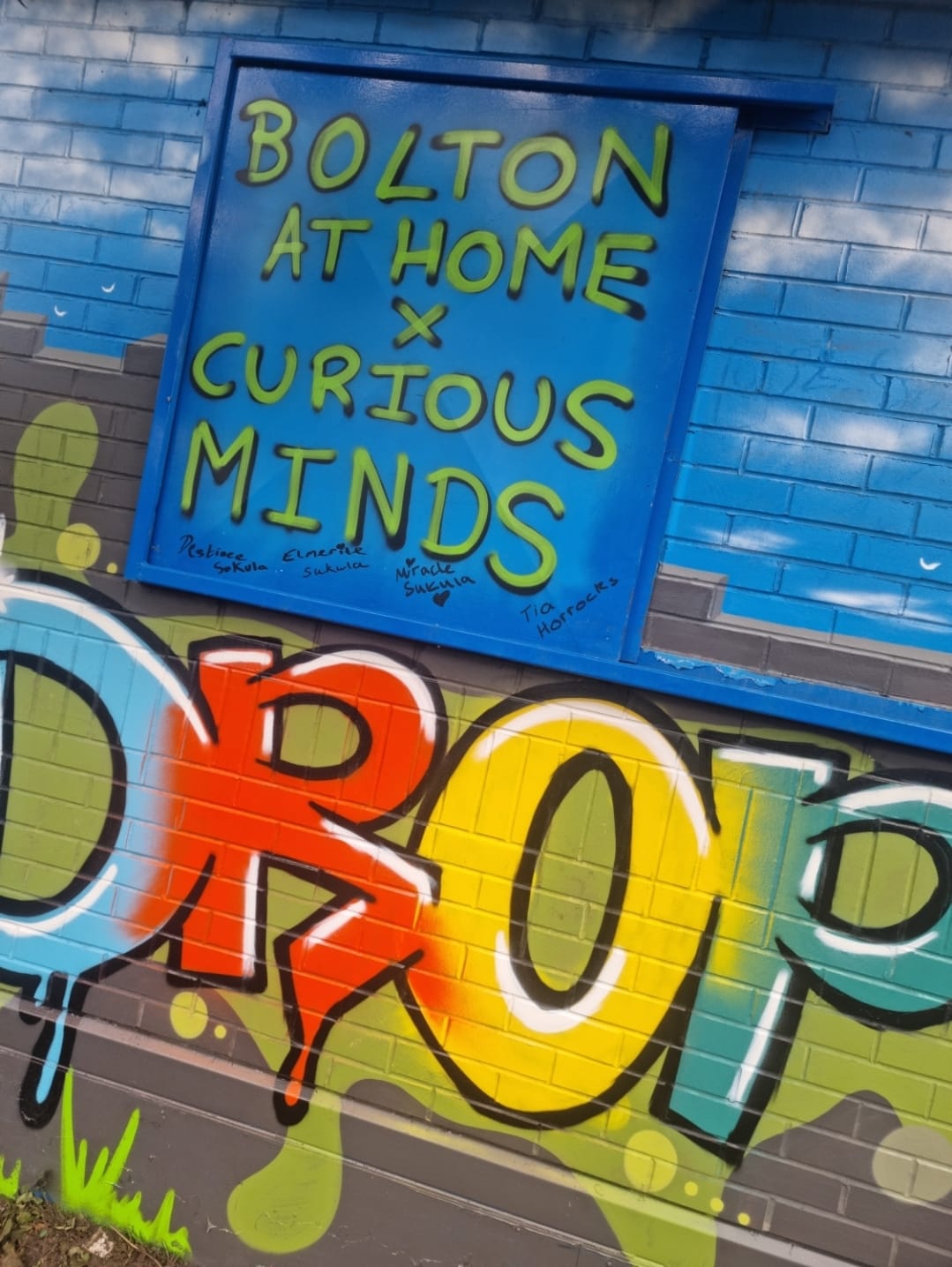 Image is a closeup of the Bolton@Home grafitti artwork. It is very colourful. A sign reads Bolton @ Home x Curious Minds