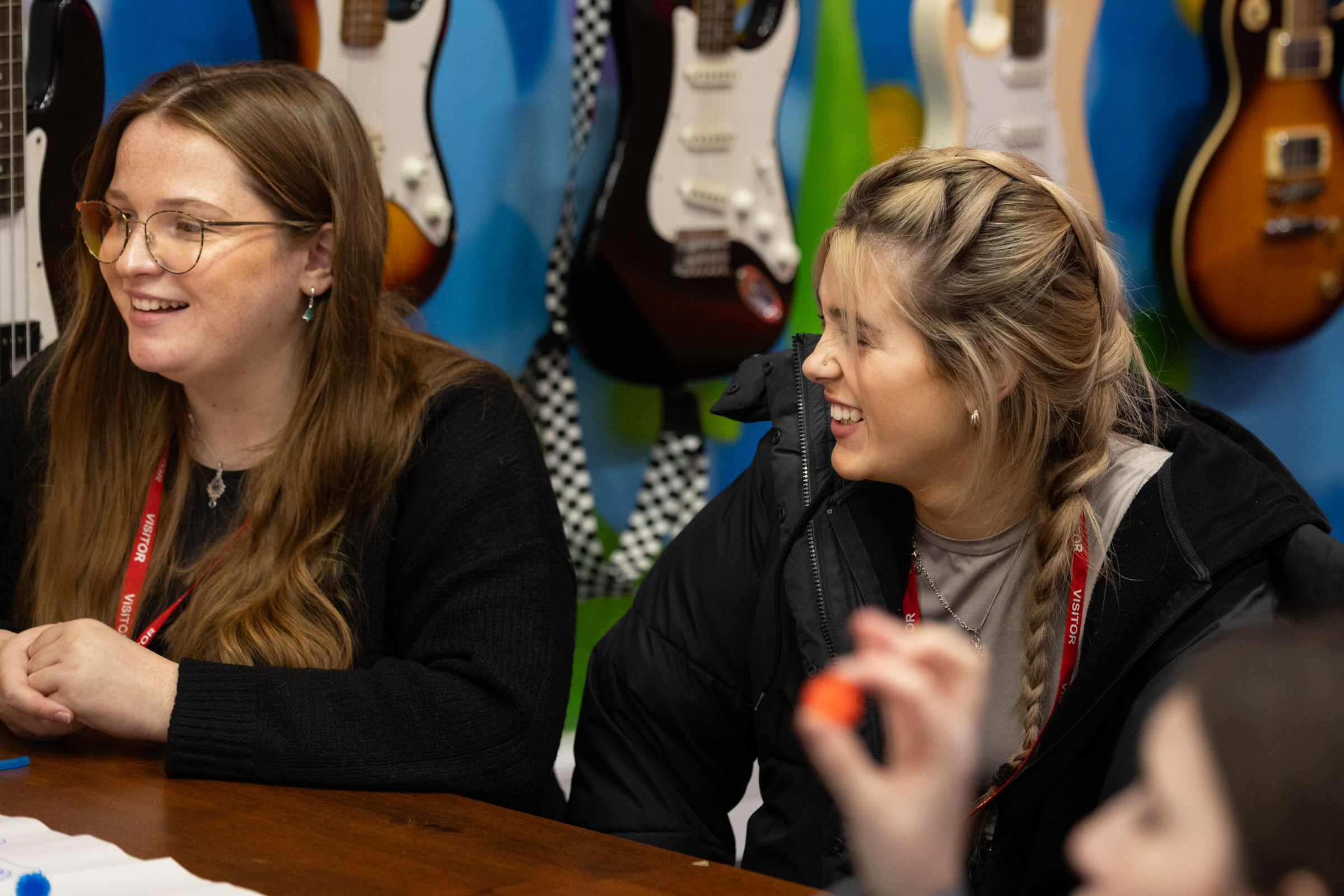 Two young females sit at a desk in front of a wall of colourful electric guitars. They are smiling and laughing.
