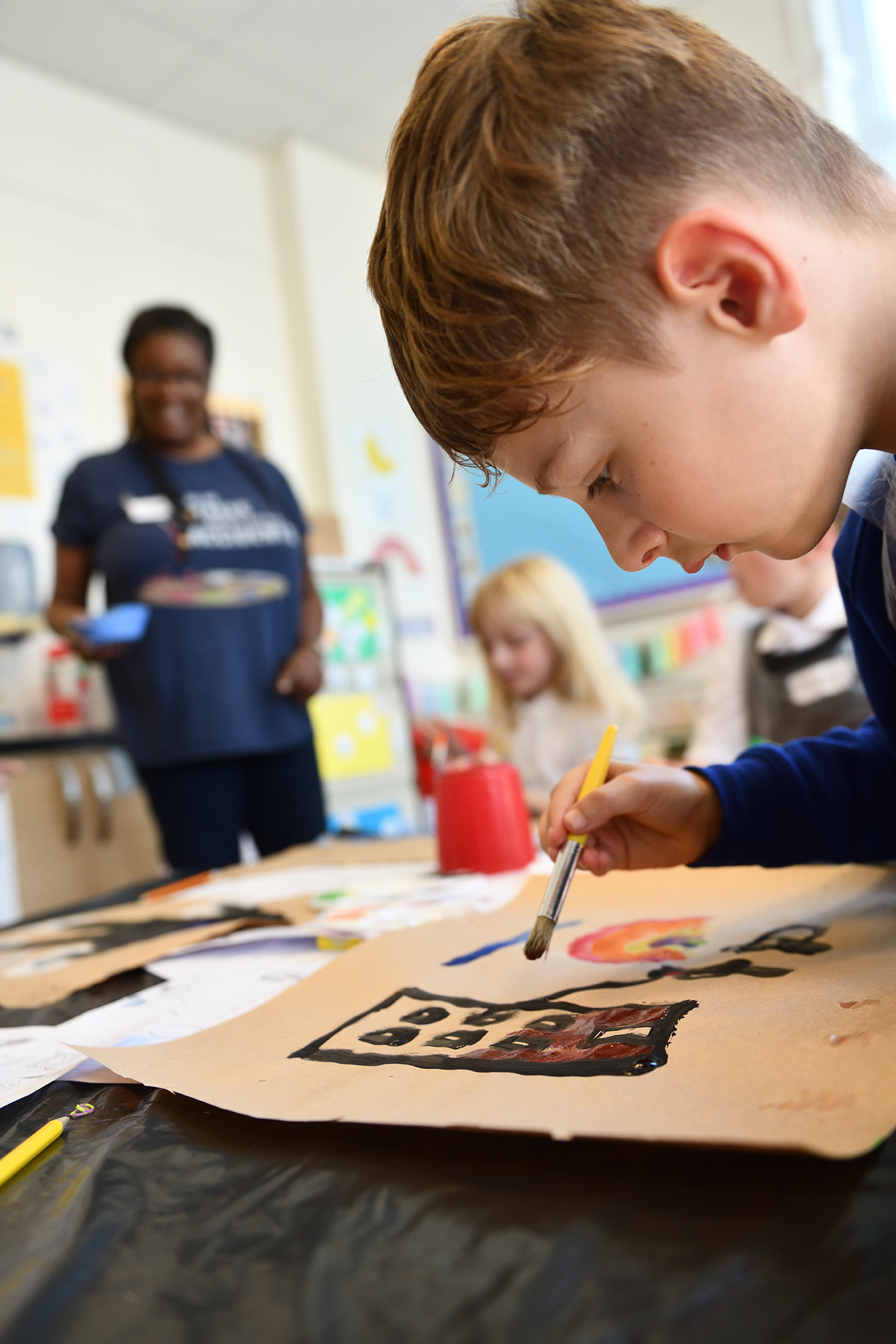 A young school pupil paints in class. His teacher can be seen in the background.