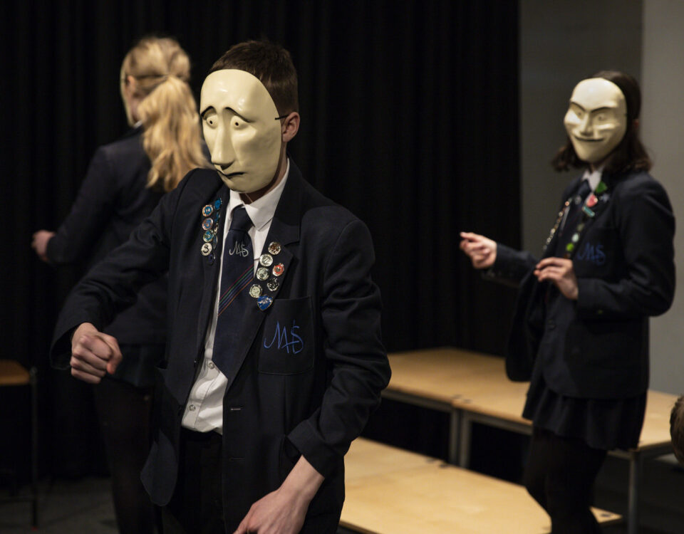 Young people in school uniform are doing a drama workshop. Two of them are using the comedy and tragedy muses masks.