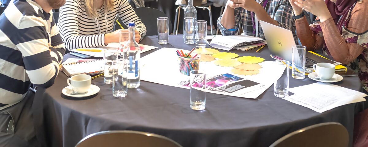 A group of people are sitting around a table, deep in discussion. There are glasses and leaflets scattered across the table.