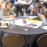 A group of people are sitting around a table, deep in discussion. There are glasses and leaflets scattered across the table.