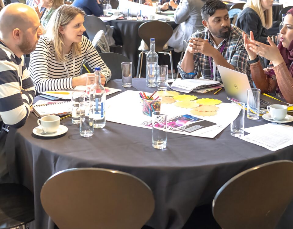 A group of people are sitting around a table, deep in discussion. There are glasses and leaflets scattered across the table.