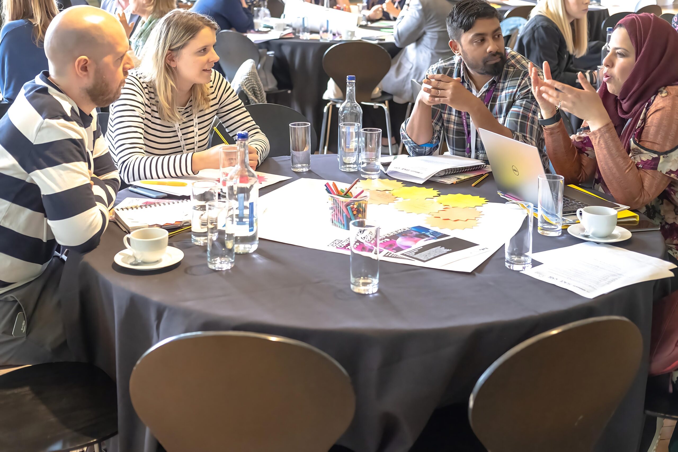 A group of people are sitting around a table, deep in discussion. There are glasses and leaflets scattered across the table.