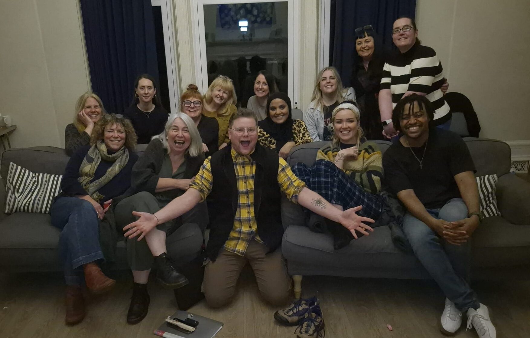 A group of 14 people are sat on and stood behind a settee. They are giving the camera big, open mouthed smiles and look excited to be together as a group.