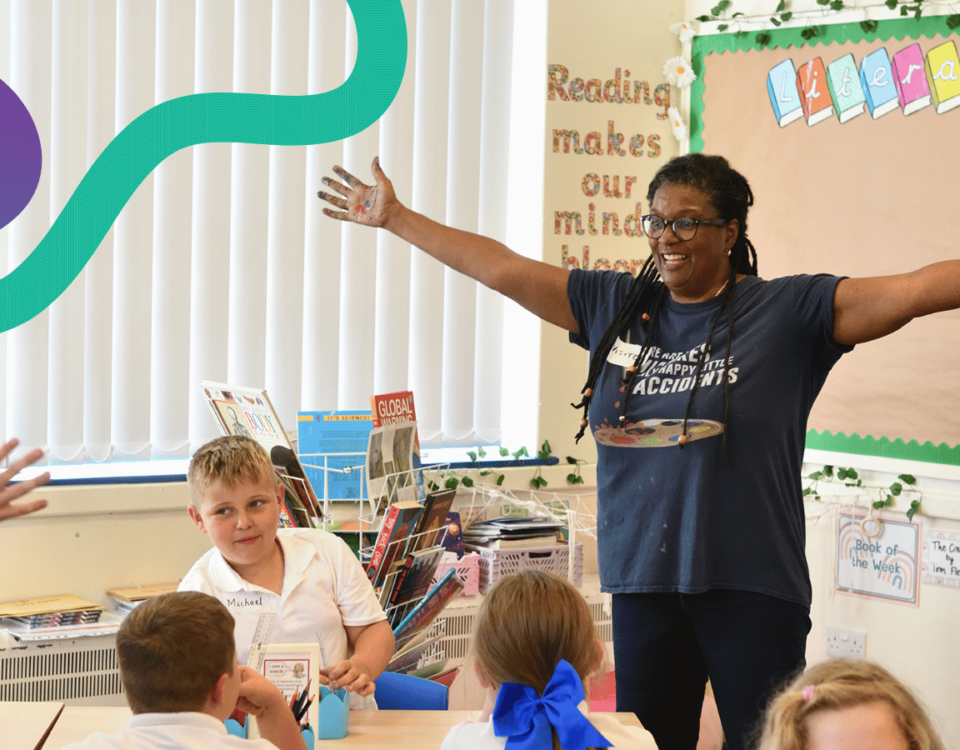 Image shows a Teaching Artist in a primary school classroom. The artist is in full flow, with arms outstretched, and a happy smile. The school pupils are engaged and enjoying their art lesson.