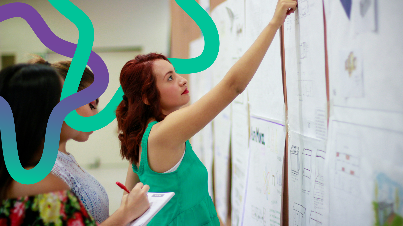 A woman with red hair stood pointing at a wall full of large pieces of paper with notes and sketches on.