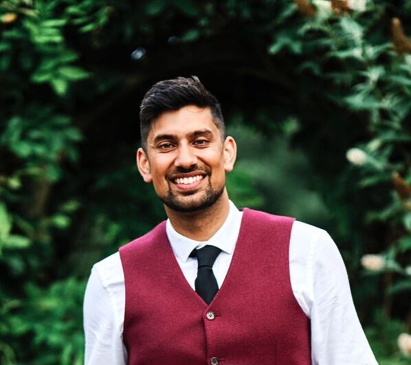 A smiling man with a beard is looking towards the camera. He's wearing a suit and is outside with a bunch of greenery.