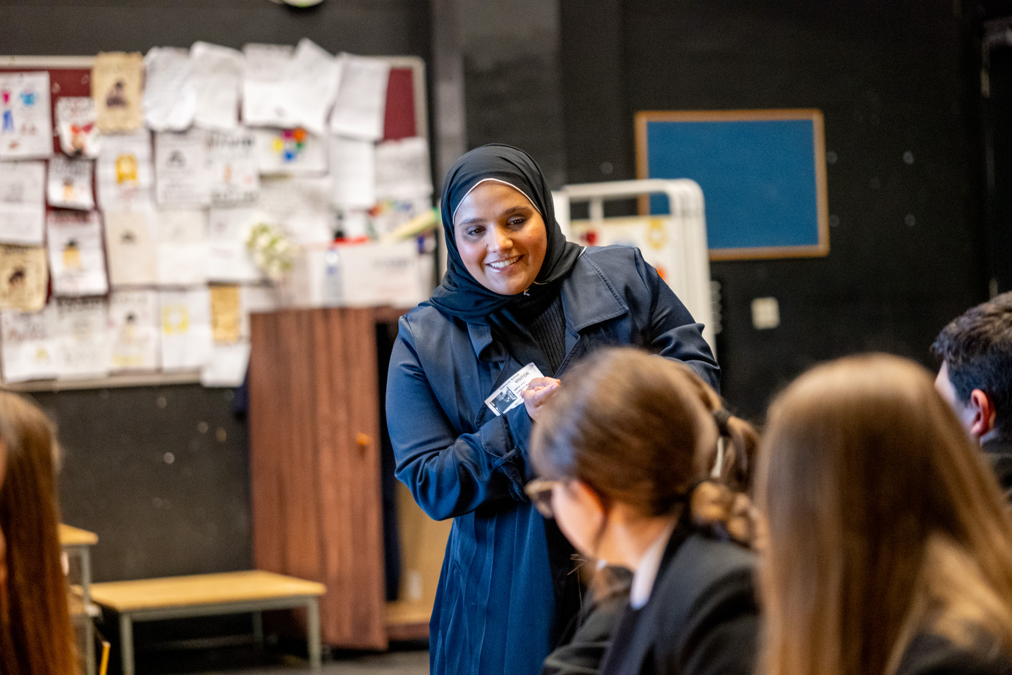 A young woman stood smiling to a couple of high school students.