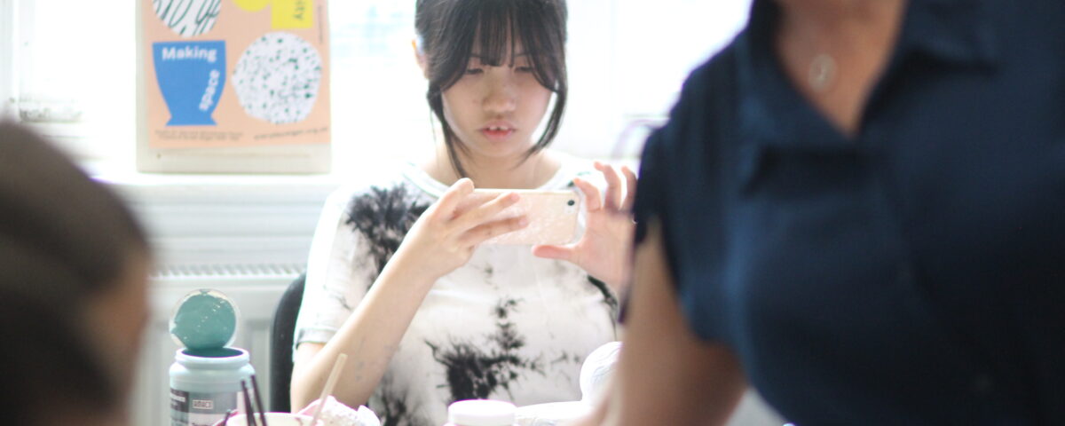A girl with dark hair sat in a pottery class taking a photo of her work.