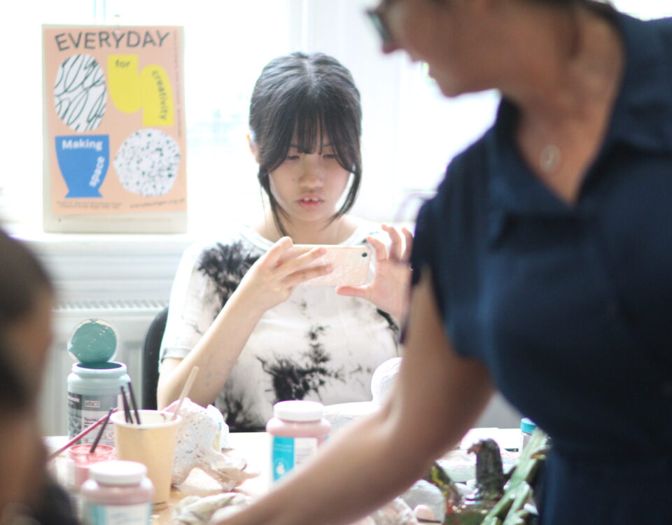 A girl with dark hair sat in a pottery class taking a photo of her work.